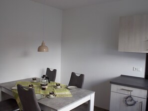 A dining area with a table set for four, a hanging pendant light, and a kitchen area with cabinets and a door. - Strandflieder by Interhome (Nessmersiel)