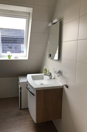 Een moderne badkamer met een wit wastafelmeubel, een spiegel en een raam met uitzicht op een gebouw.