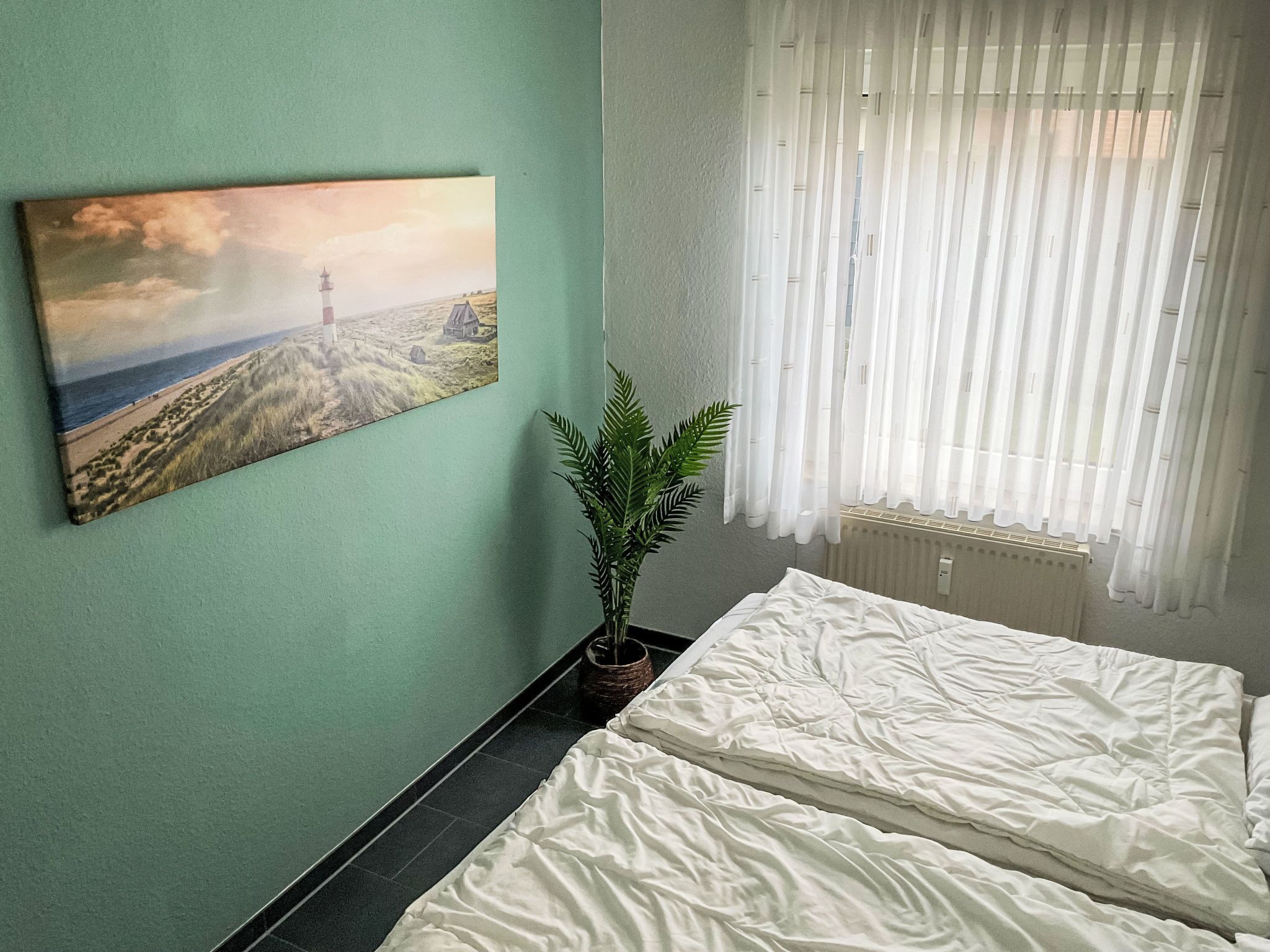 A room with a bed, a potted plant, a painting, and a window with sheer curtains.