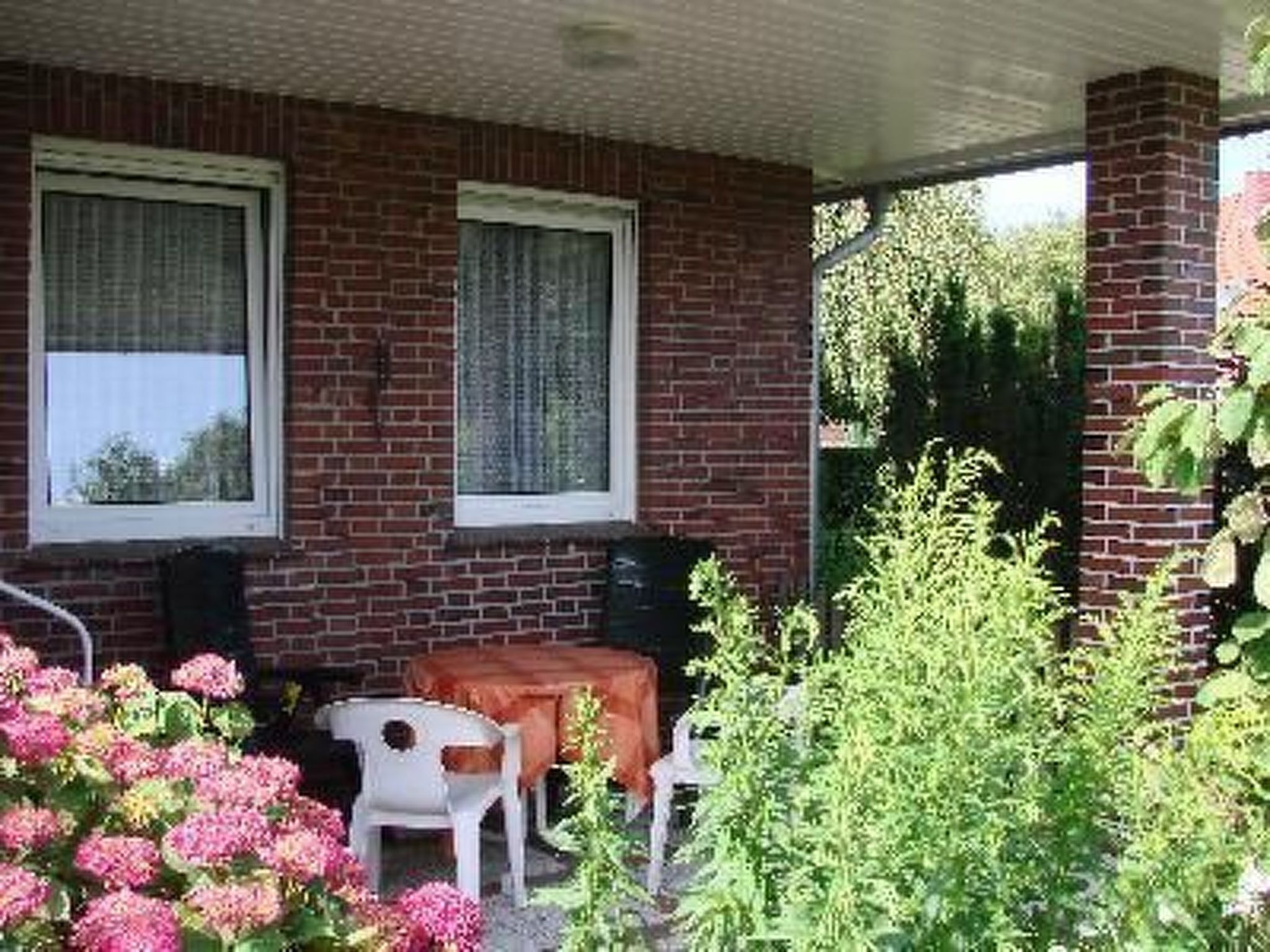 A covered patio area with a brick wall, two windows with curtains, a small table with chairs, and a variety of plants.