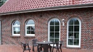 Ein Backsteinhaus mit Ziegeldach, drei Rundbogenfenstern und einer Terrasse mit Tisch und Stühlen.