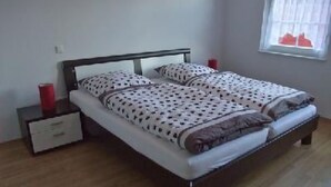 Ein Bett mit gemustertem Überwurf, ein Nachttisch mit einem roten Gegenstand und ein Fenster mit Jalousien.