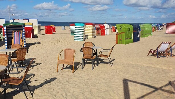 Ein Strand mit Strandkörben, Korbstühlen und Tischen.