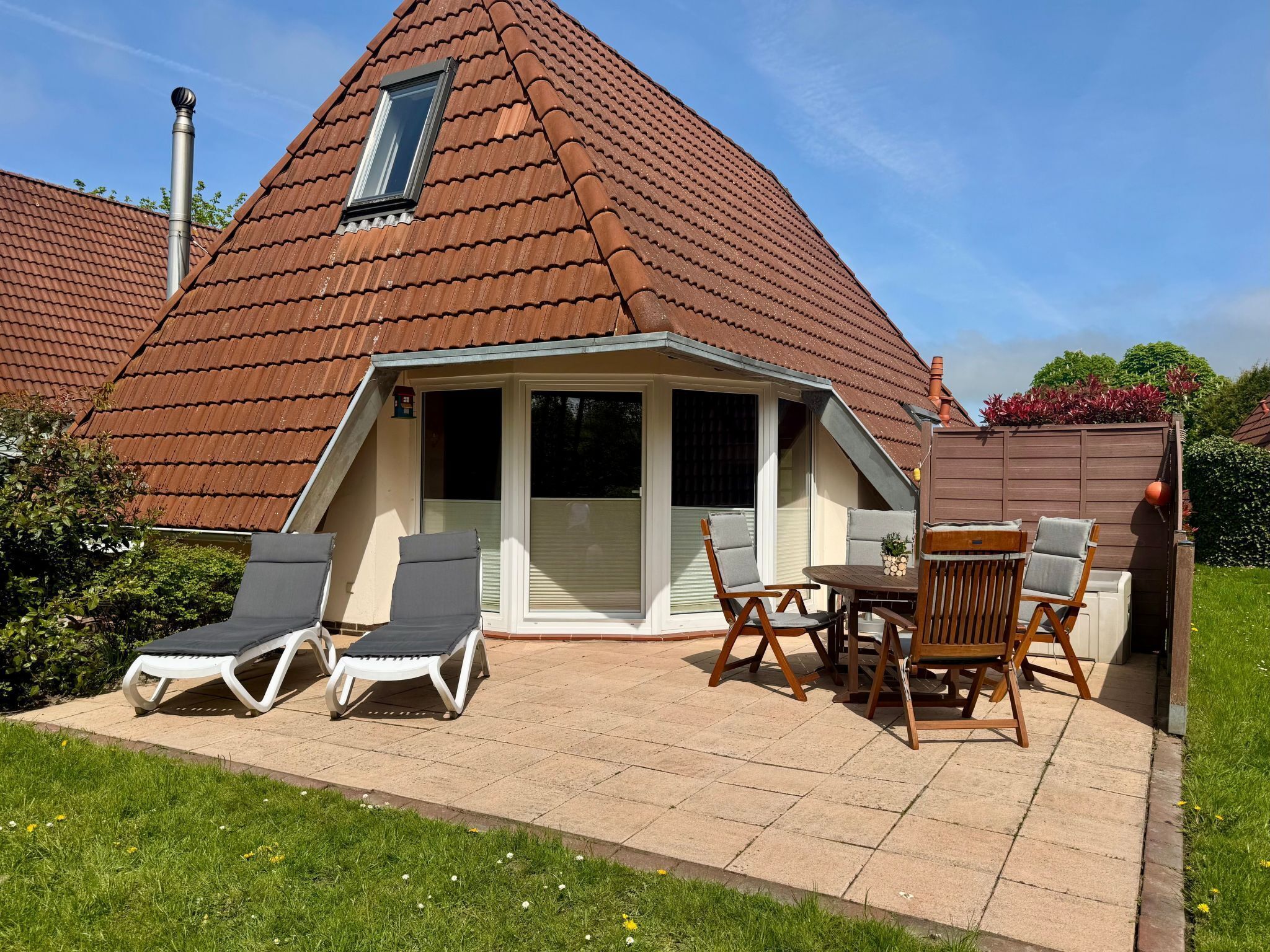 Ein Haus mit geneigtem Dach, einer überdachten Terrasse mit Liegestühlen und einem Tisch sowie einem kleinen Garten.