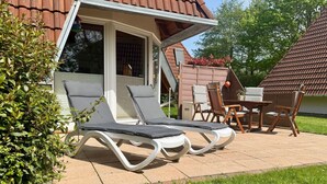 Een terras met twee loungestoelen en een tafel, omgeven door een tuin en een huis met een pannendak.