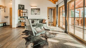 Een ruime woonkamer met een hoge plafond, houten balken, een groot raam met gordijnen, een bank, een bijzettafeltje en een vloerkleed.
