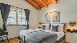 Een slaapkamer met een houten plafond, een groot bed met grijze beddengoed, een stoel en kunst aan de muur.