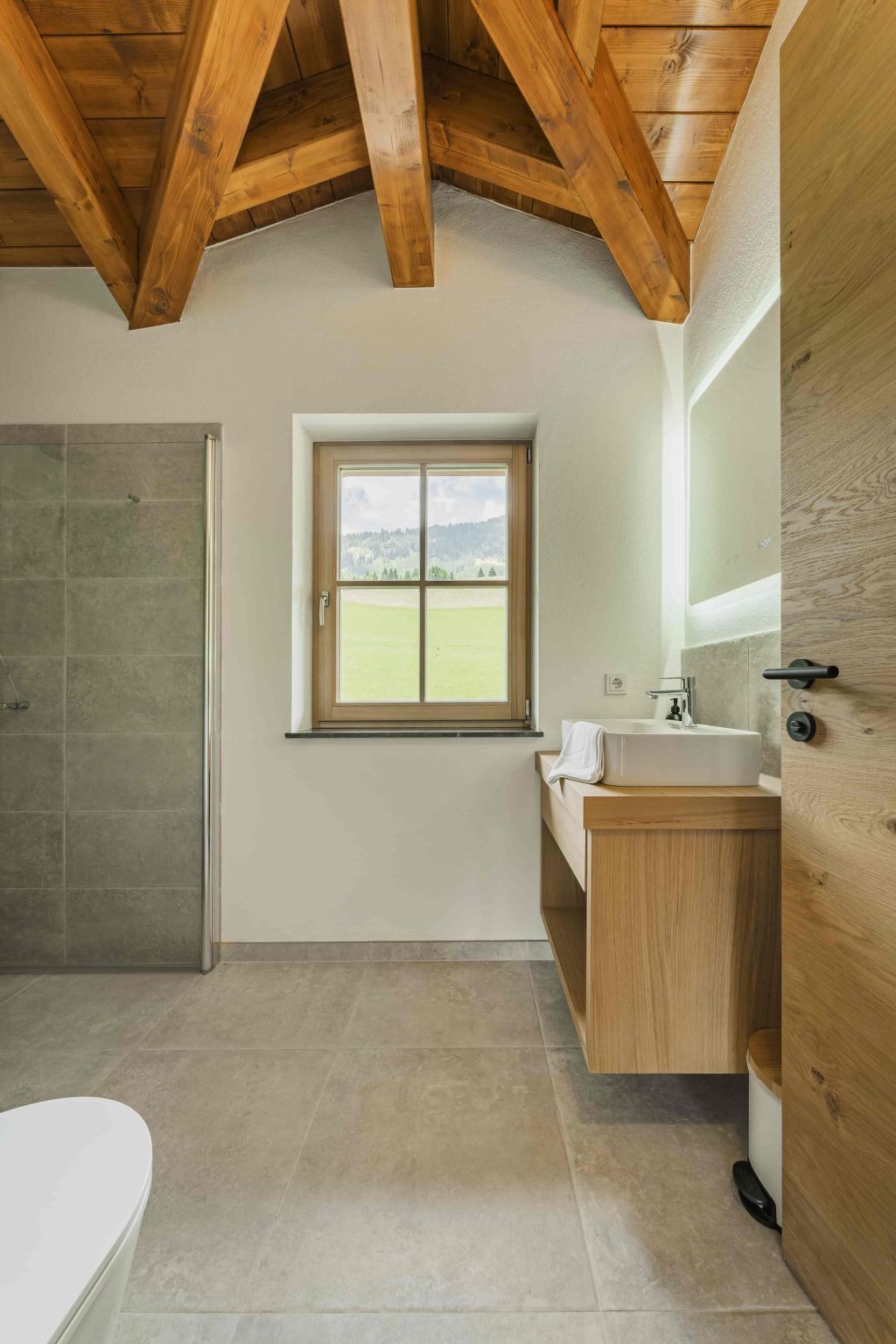 A bathroom with a wooden ceiling, a window with a view of a green field, a sink, and a toilet.