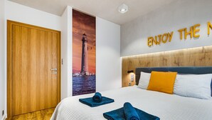 Ett hotellrum med en trädörr, en stor säng med blått och vitt sänglinne, en affisch med en fyr och en väggskylt med texten ”NJUT AV N”.