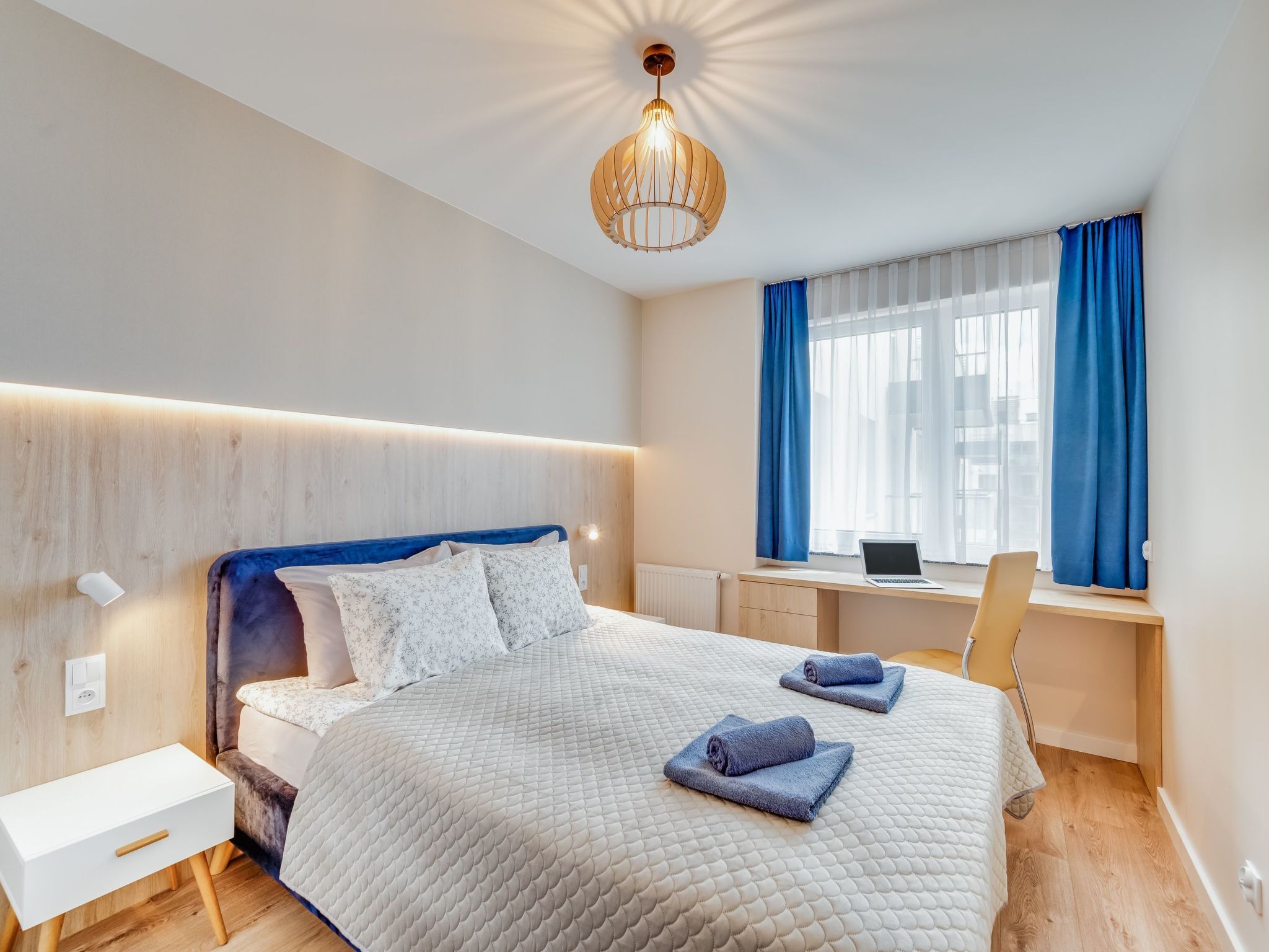 A modern bedroom with a bed, bedside table, desk, and laptop.