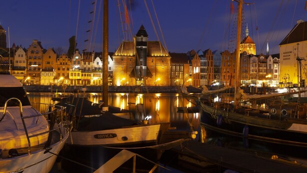 Ein Hafen im Abenddämmerung mit vertäuten Booten, ein markantes Gebäude mit einem Turm, und ein Gebäude mit der Aufschrift "Narodowe Muzeum Morskie".