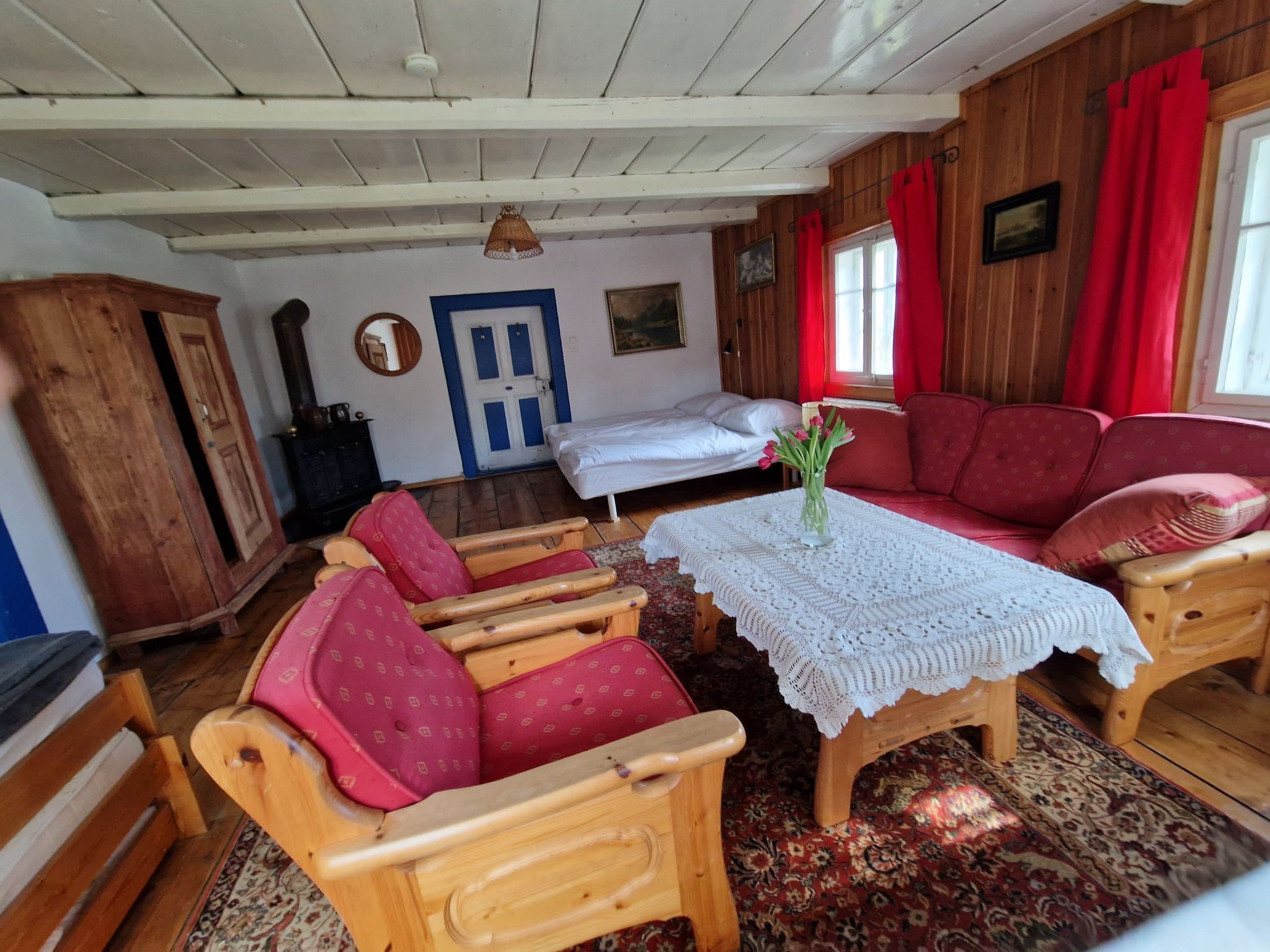 Habitació acollidora amb sostre de fusta, un sofà vermell, una taula de fusta amb estovall i un llit amb roba de llit blanca.