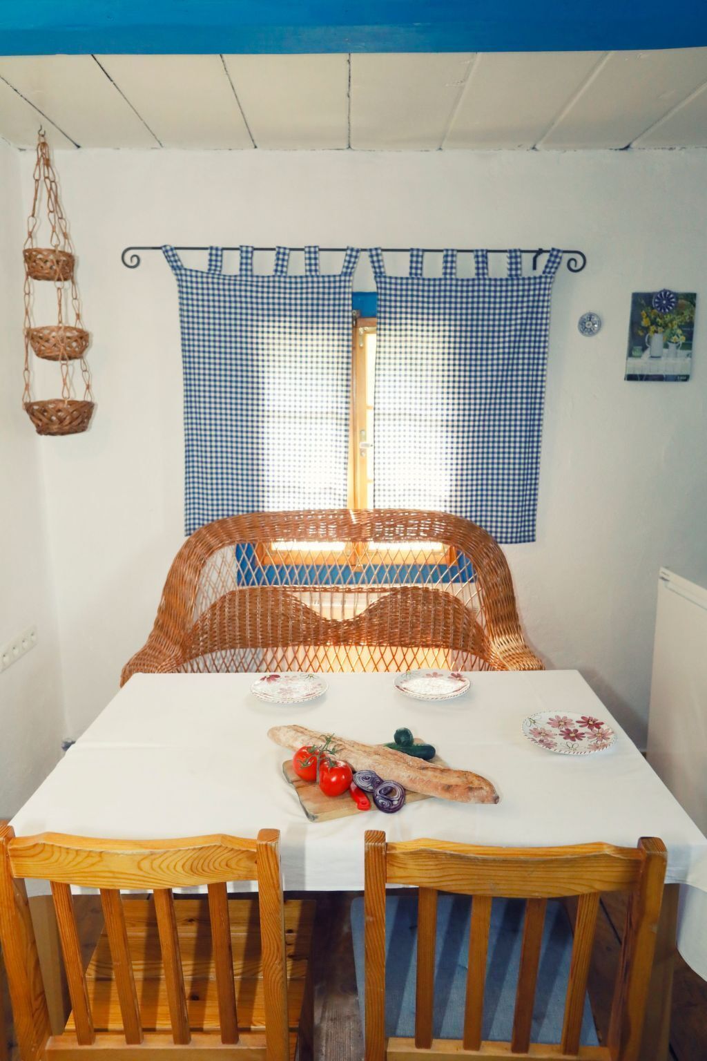 Un comedor con una mesa blanca, una silla de mimbre y una cortina a cuadros.