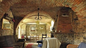 Ein gemütliches Zimmer mit Steinwänden, einer Chandelierleuchtre, einem Kamin und einem Essbereich, der durch einen Rundbogen sichtbar ist.