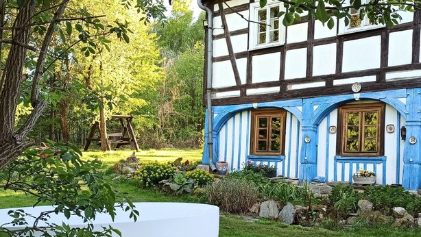 Ein traditionelles Fachwerkhäuschen mit gepflegtem Garten, einer Schaukel und einem großen weißen Blumentopf im Vordergrund.