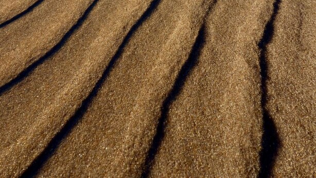 En strand med vågformade sandmönster och ett stilla vatten som speglar himlen.