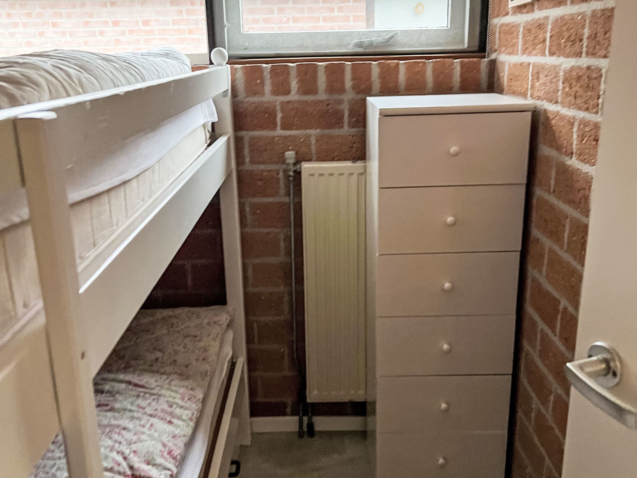Een stapelbed met een raam erboven, een radiator en een dressoir in een kamer met bakstenen muren.