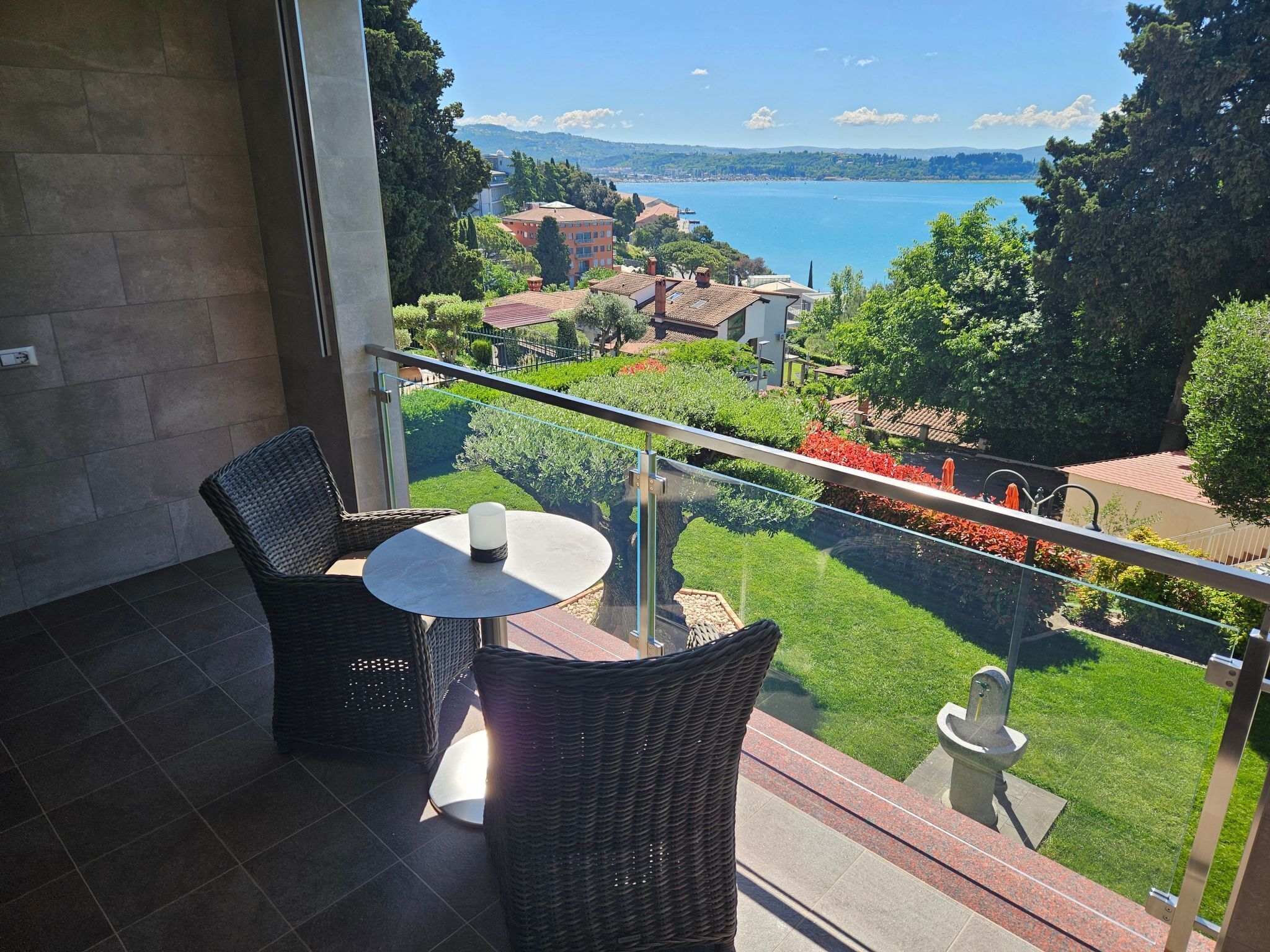Un balcon agrémenté de fauteuils et d’une table en osier, donnant sur un jardin aménagé et un lac, avec des bâtiments visibles au loin.