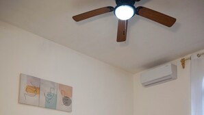 Un dormitori amb un llit, un ventilador de sostre, un aparell d'aire condicionat i una obra d'art penjada a la paret.