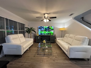 A living room with two white leather sofas, a glass coffee table, a ceiling fan, a flat-screen TV, and a lamp. - Lakeside Getaway Near AT&T Stadium, Globe Life, Six Flags & Epic Waters (Grand Prairie)