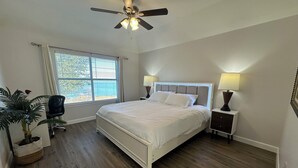 Une chambre à coucher comprenant un lit, des lampes de chevet, un ventilateur de plafond, une fenêtre avec des stores, une chaise et une plante en pot.