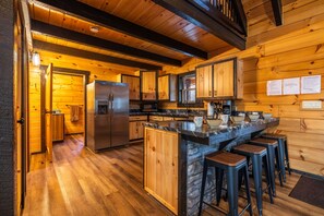 A log cabin interior with a kitchen featuring wooden cabinets, a stone island with bar stools, and a stainless steel refrigerator. - Schilling Hill (New Philadelphia)