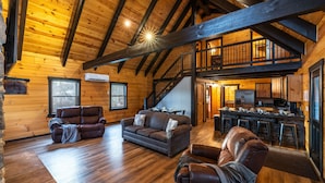 A spacious log cabin interior with a wooden ceiling, a living area with leather sofas, a kitchen with stone countertops, and a staircase leading to an upper level. - Schilling Hill (New Philadelphia)