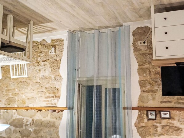 Una habitación con paredes de piedra, techo de madera, cómoda blanca, ventana con cortina y una pequeña estantería con una lámpara.