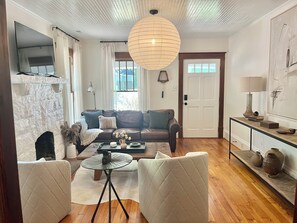 A living room with a brown leather sofa, a round table, and a white armchair. There is a fireplace, a TV mounted on the wall, and a door with a window. - The Vintage Vibe- Five Points, sleeps 6, pets ok (Raleigh)