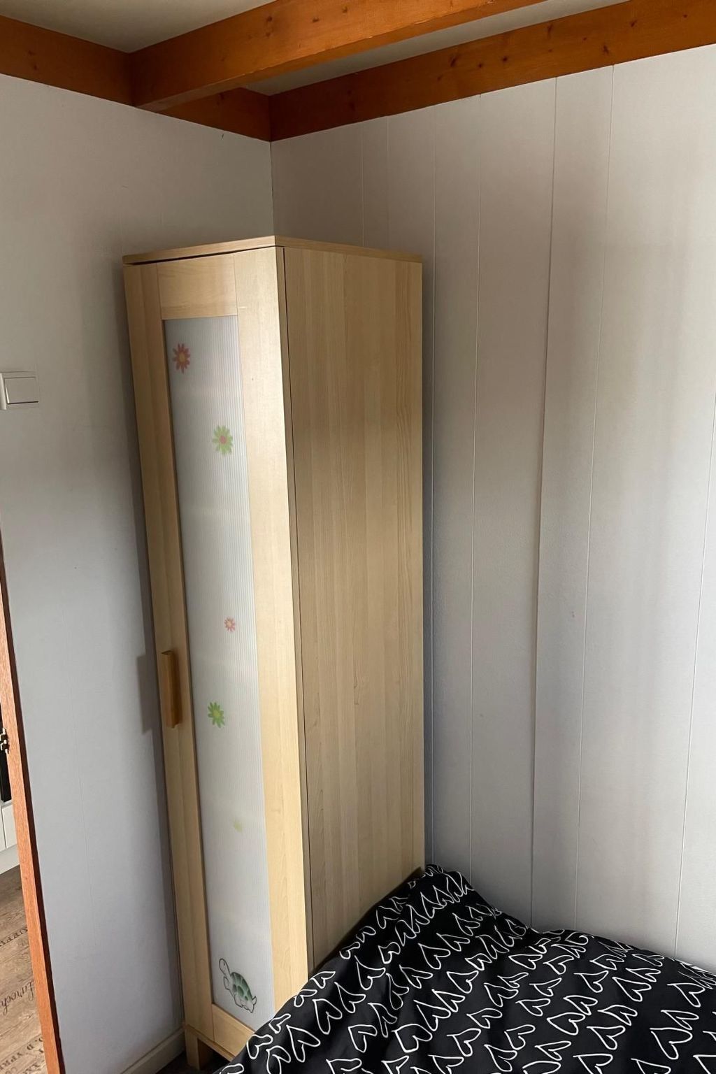 A room with a wooden wardrobe, a bed with a patterned bedspread, and white walls.
