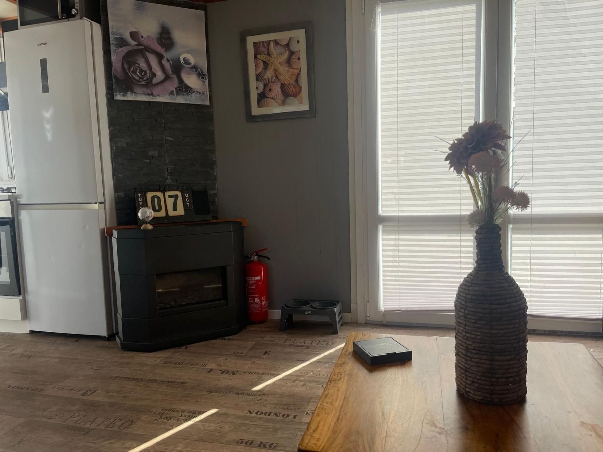 A kitchen with a refrigerator, a fireplace, a fire extinguisher, and a vase with flowers on a wooden table.