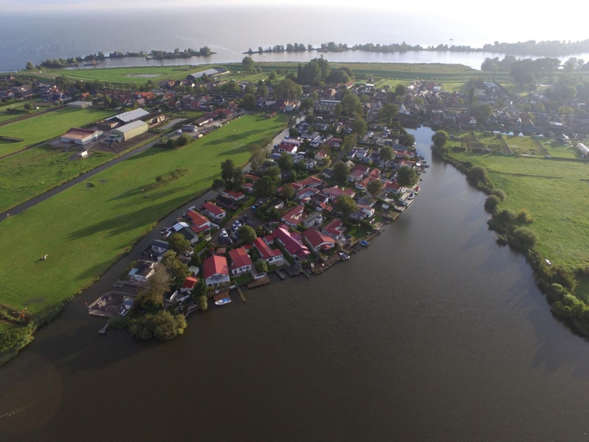 An aerial view of a riverside village with houses along the water, green fields, and a distant shoreline.