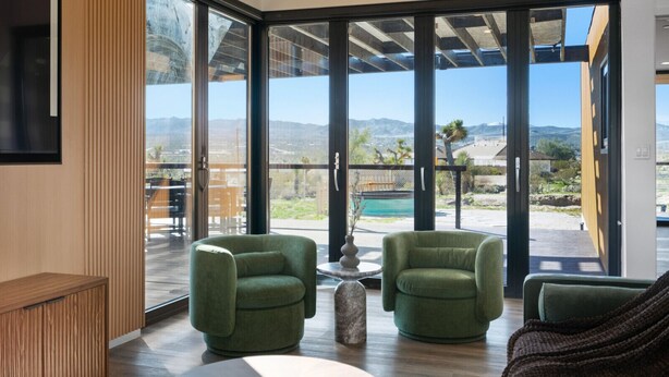 A spacious room with large windows offering a view of mountains and a pool. There are two green armchairs and a wooden bench. - Oasi Espresso|AMAZING mountain views|Cantilevered house w/cowboy pool (Yucca Valley)