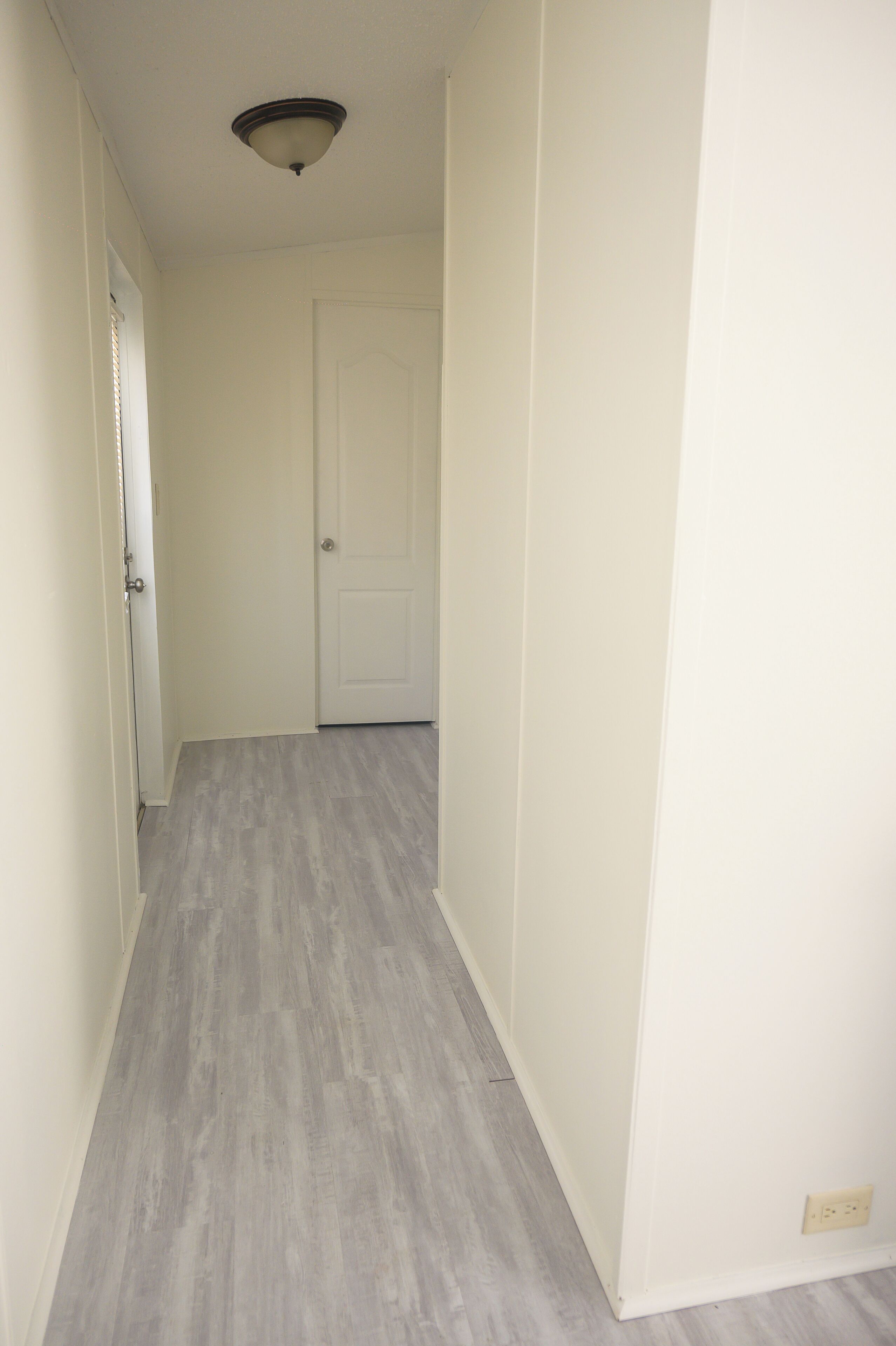 Un couloir étroit, avec une porte blanche au fond, un parquet en bois de couleur claire et un luminaire au plafond.