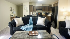 A living room with a black sofa, a coffee table with a board game, and a kitchen in the background. - 3BR FOREST NEST in Columbus, GA (Columbus)