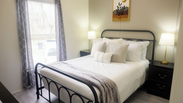 A neatly made bed with white linens and a gray throw blanket, a bedside table with a lamp, and a wall-mounted artwork. - 3BR FOREST NEST in Columbus, GA (Columbus)