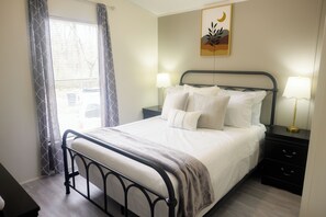 A neatly made bed with white linens and a gray throw blanket, a bedside table with a lamp, and a wall-mounted artwork. - 3BR FOREST NEST in Columbus, GA (Columbus)