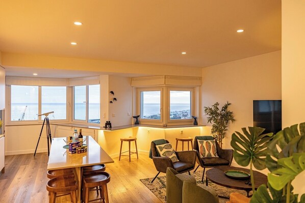 Uma sala de estar moderna com área de jantar, janelas amplas com vista para o mar e um telescópio em um tripé.