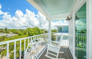 A covered porch with a white railing, a rocking chair, and a view of a green landscape. - Siesta Key Luxe w/ Pool, Rooftop & Steps to Beach (Siesta Key)