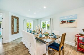 A dining room with a wooden table set for four, white chairs, and a painting of boats on the wall.