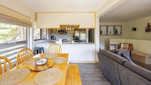 Una cucina moderna con isola centrale, tavolo da pranzo in legno con sottopiatti intrecciati e un divano grigio in zona giorno con vista sull'esterno.