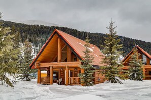 Une cabane en rondins avec un toit rouge, entourée de terre et d’arbres recouverts de neige, avec une chaîne de montagnes en arrière-plan.