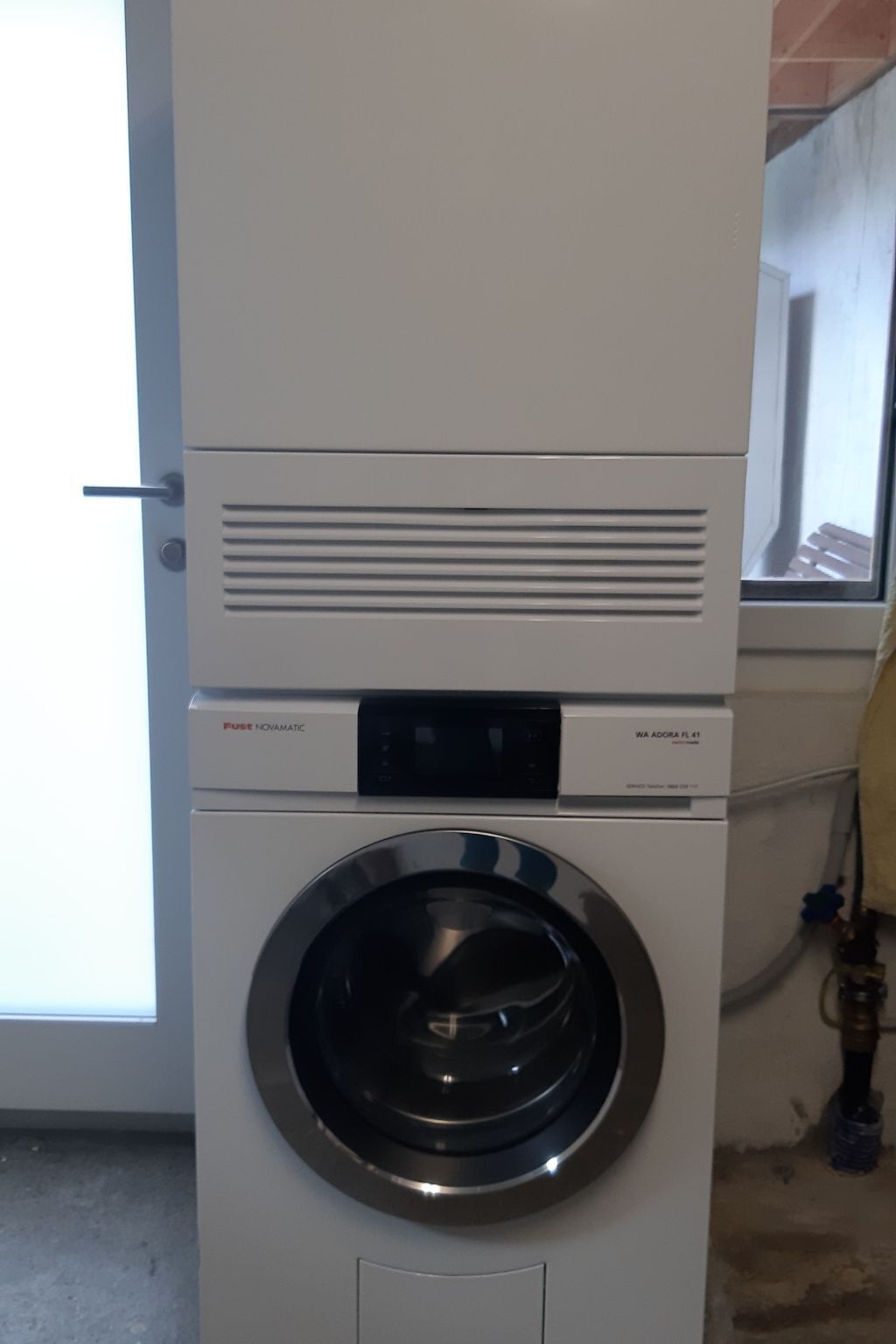 A front-loading washing machine with a digital control panel, located in a laundry room with a window and a door.