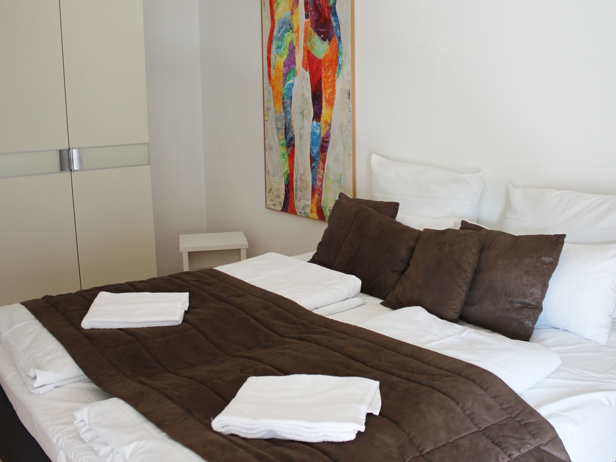 Un letto rifatto con cura, con una coperta marrone e lenzuola bianche, un comodino bianco e un quadro astratto dai colori vivaci appeso alla parete.