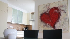 Een moderne keuken met witte kasten, een koffieapparaat en een eettafel met twee stoelen. Aan de muur hangt een groot abstract schilderij van een rood hart.