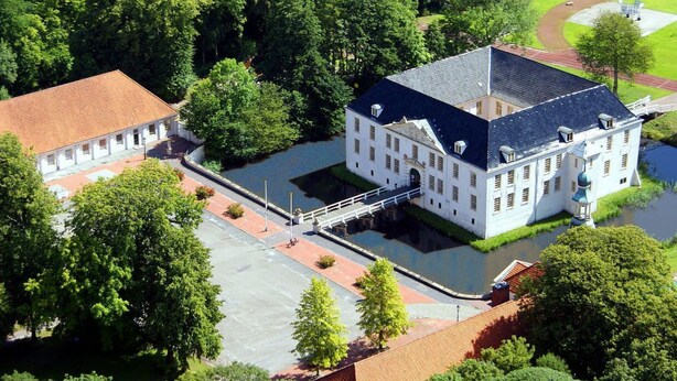 Un grand bâtiment blanc, surmonté d’un toit sombre, entouré d’une végétation luxuriante et bordé d’un canal.