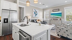 Cocina moderna con refrigerador de acero inoxidable, muebles blancos y una isla grande con pileta. Hay un comedor con una mesa blanca y sofá estampado, y un ventanal con vista a la playa.
