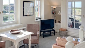 Un salon chaleureux avec une table blanche, un fauteuil marron, un canapé bleu et un téléviseur. Il y a des fenêtres donnant sur la verdure et une lampe dans un coin.