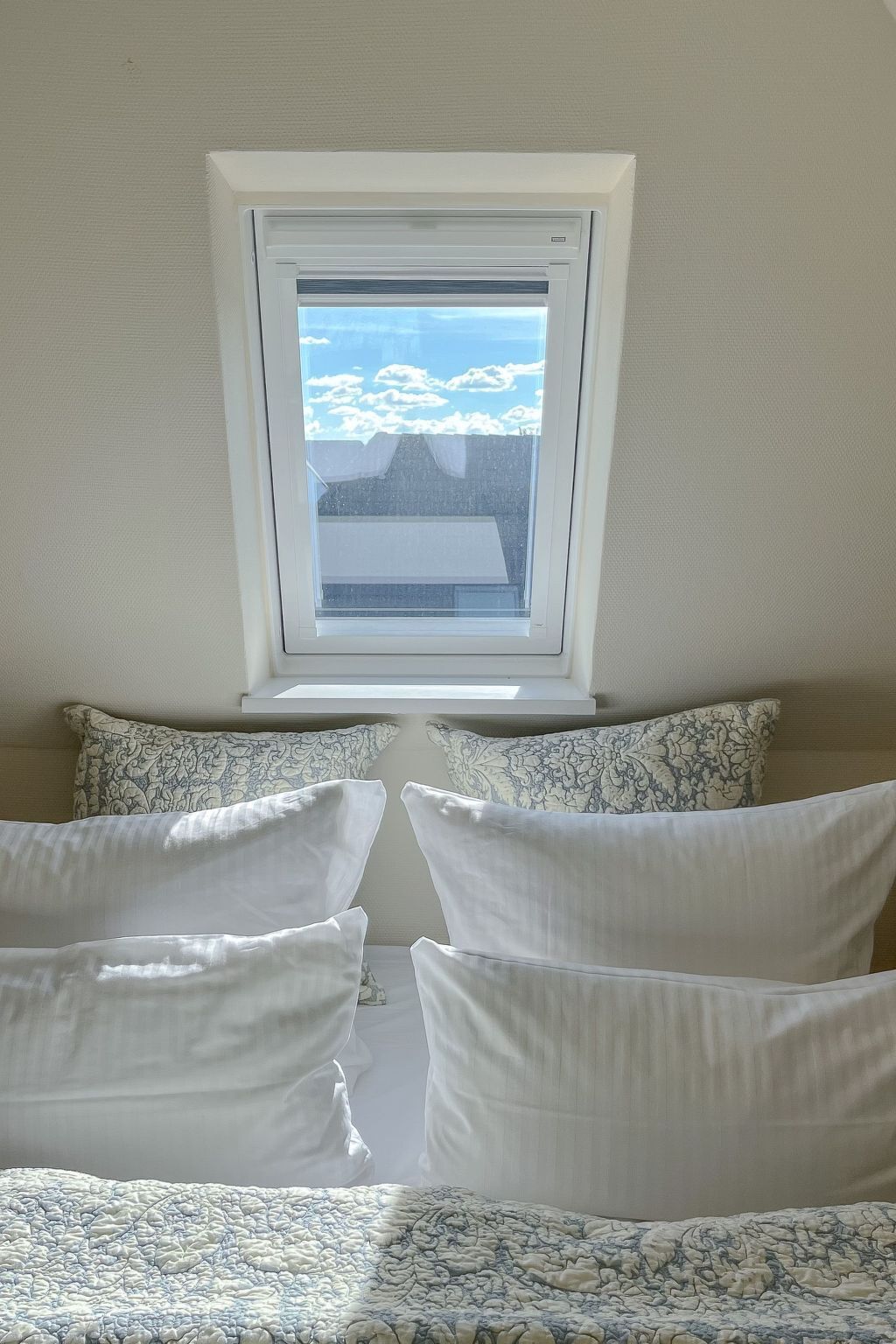 Une chambre avec une lucarne, un lit recouvert d’une literie à motifs et une vue sur le ciel et les nuages.