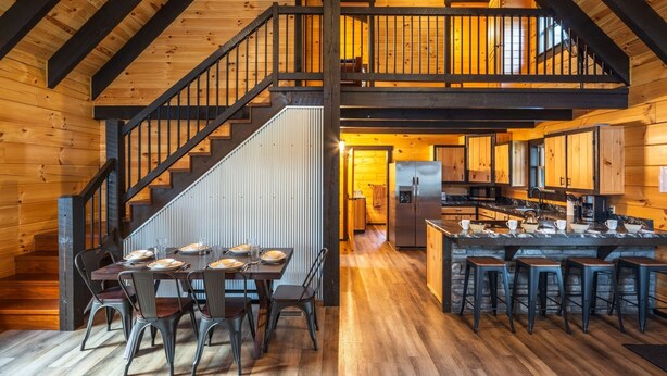 Une cuisine spacieuse avec des poutres apparentes, un îlot central, des tabourets de bar et un escalier menant à une mezzanine.